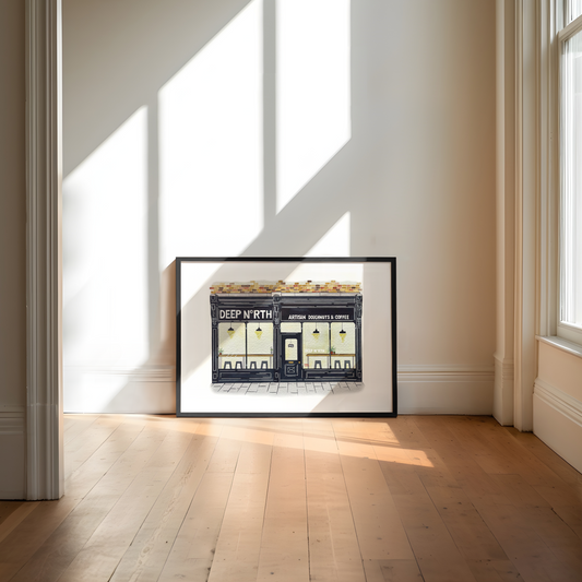 A framed art print depicting a shop storefront with the text 'Deep North" is leaning against a wall on a light wood floor in a sunlit room.
