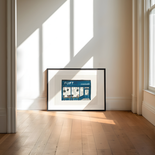 Framed artwork of a pizza restaurant on a wall in a bright room with sunlight streaming in.
