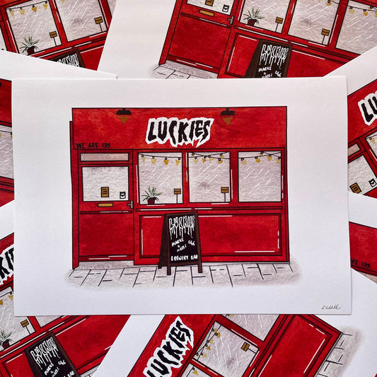 A white art print featuring a hand-drawn illustration of a red shop front with the word 'LUCKIES' in bold white letters, surrounded by multiple overlapping copies of the same print.