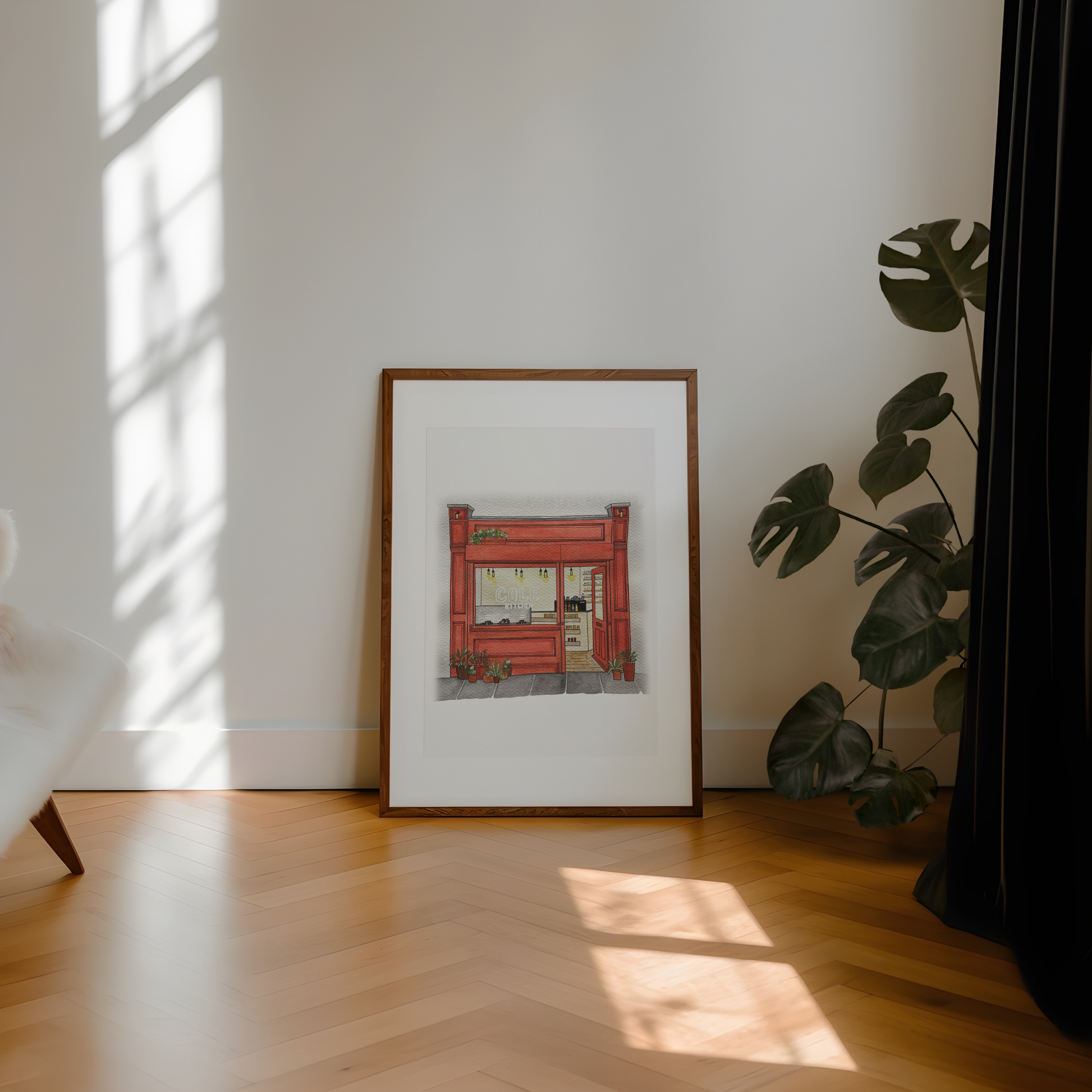 Framed artwork on a wall with a plant and sunlight casting shadows on a wooden floor.