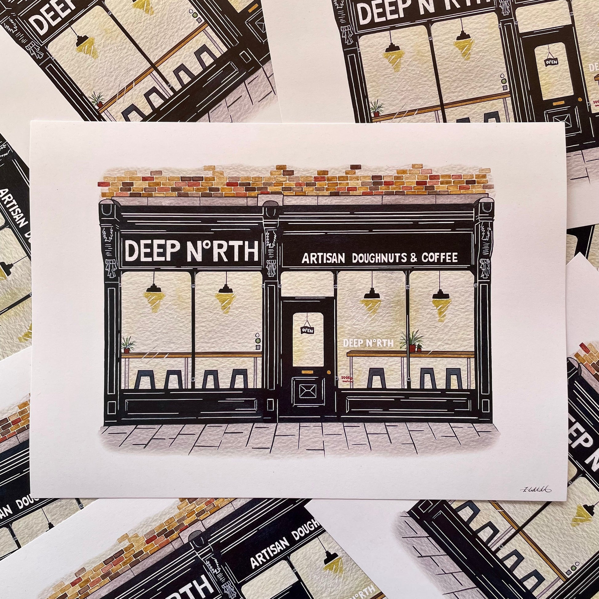 A printed illustration of a storefront reading 'DEEP NORTH' and 'ARTISAN DOUGHNUTS & COFFEE' is shown on a white sheet, surrounded by multiple copies of the same print.