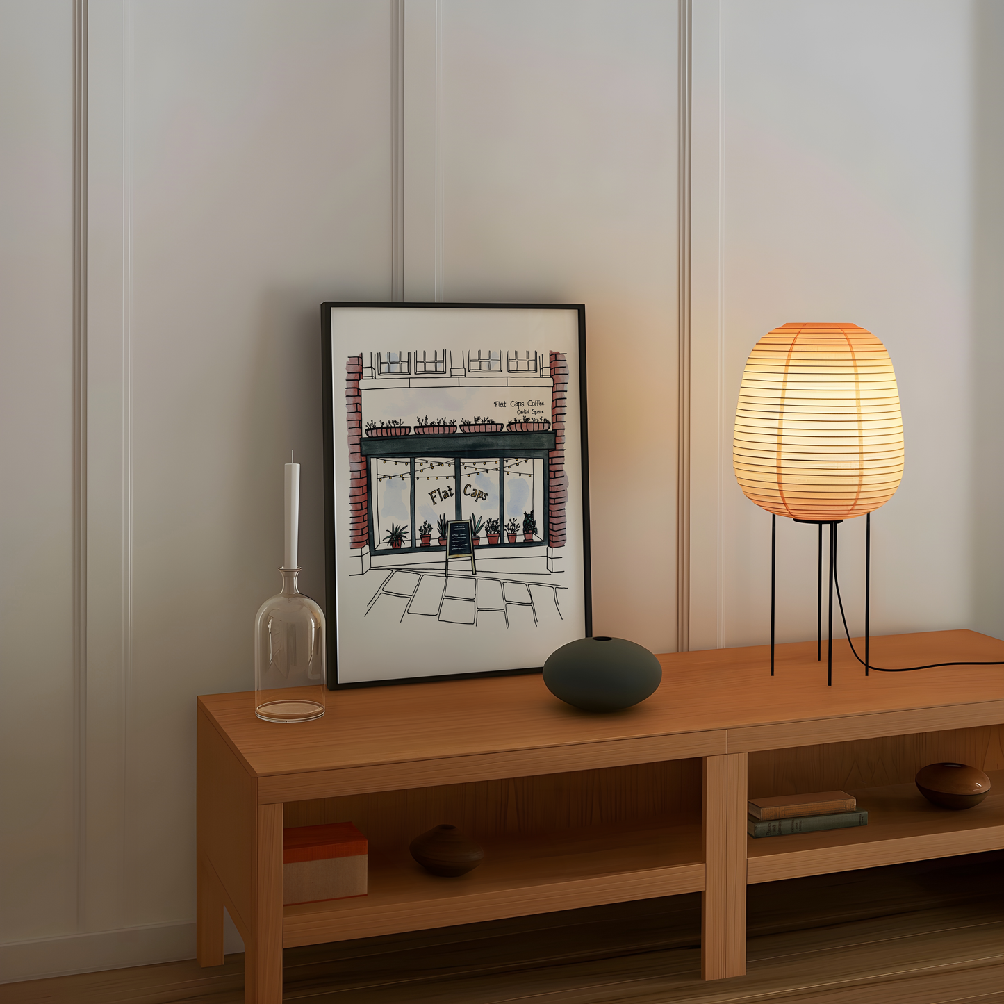 Wooden shelf with a framed picture of Flat Caps coffee shop, candle, vase, and lamp against a white wall.