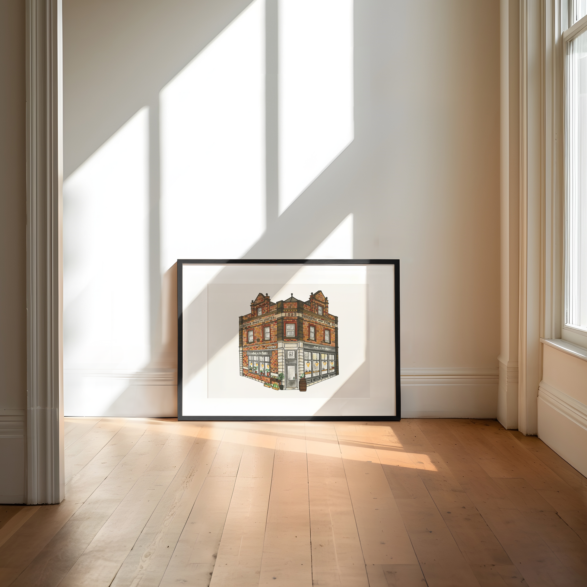 Framed artwork of a building on a wall in a bright room with sunlight streaming through windows.