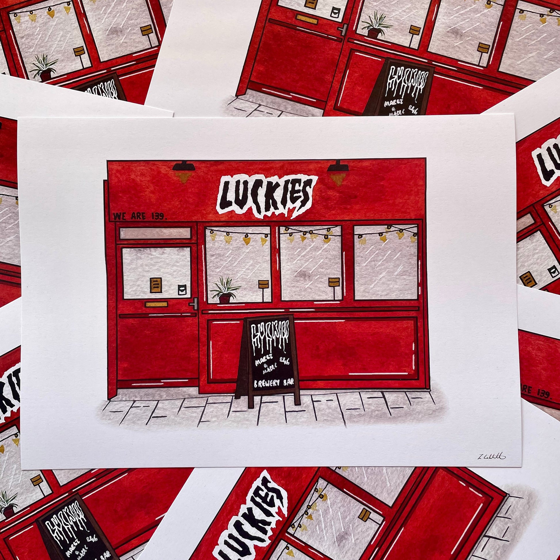 A white art print featuring a hand-drawn illustration of a red shop front with the word 'LUCKIES' in bold white letters, surrounded by multiple overlapping copies of the same print.