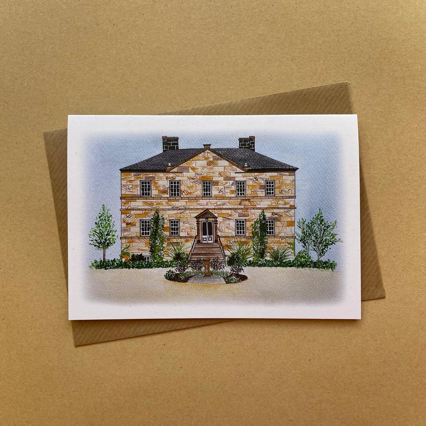 Greeting card with an illustration of Newton Hall wedding venue illustration displayed on a brown background