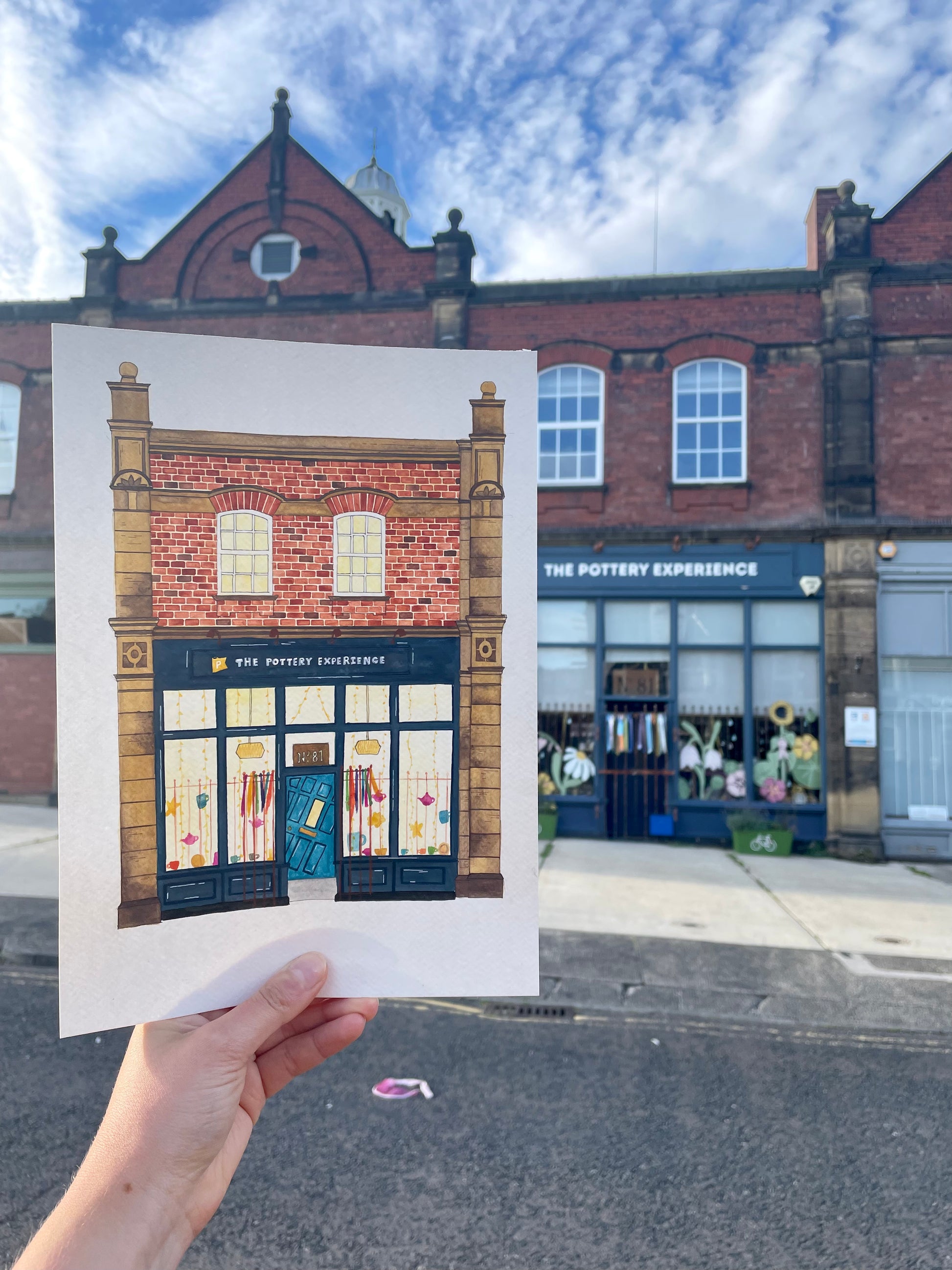 Hand holding a print of The Pottery Experience in front of the actual building.