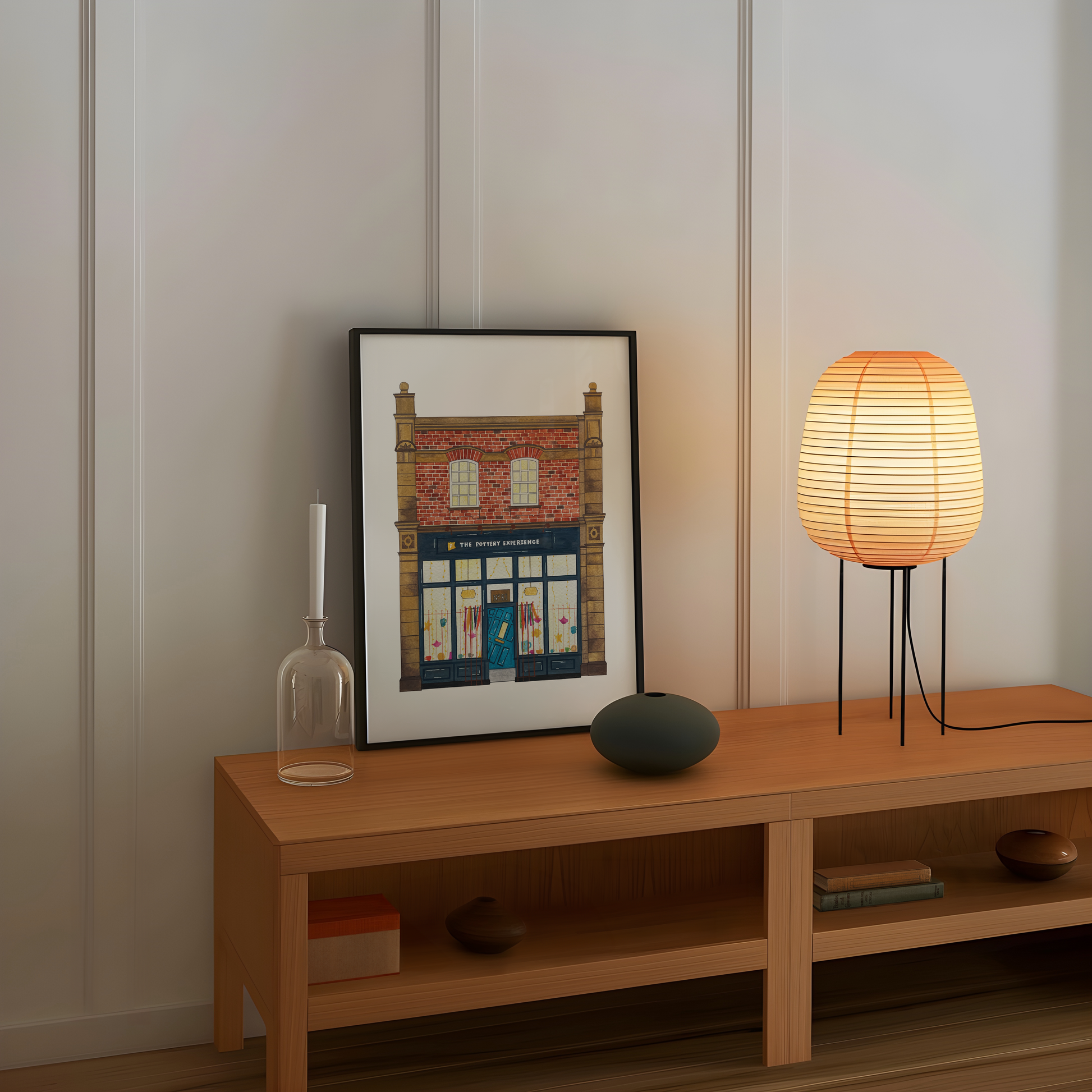 Wooden shelf with a framed picture of The Pottery Experience Jesmond, candle, vase, and lamp against a white wall.