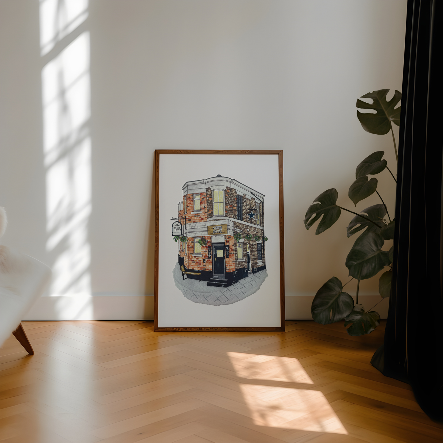 A framed art print of a street corner pub, leaning against a white wall on a light wood herringbone floor next to a potted monstera plant.