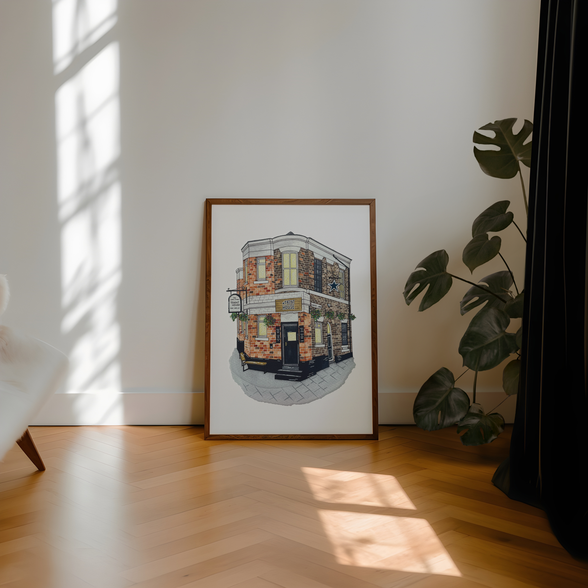 A framed art print of a street corner pub, leaning against a white wall on a light wood herringbone floor next to a potted monstera plant.