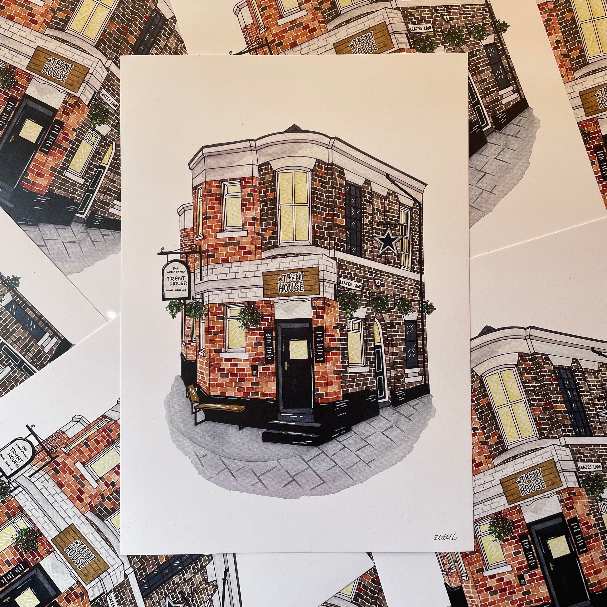 An art print featuring a detailed illustration of a brick pub building, displayed on top of several matching prints.