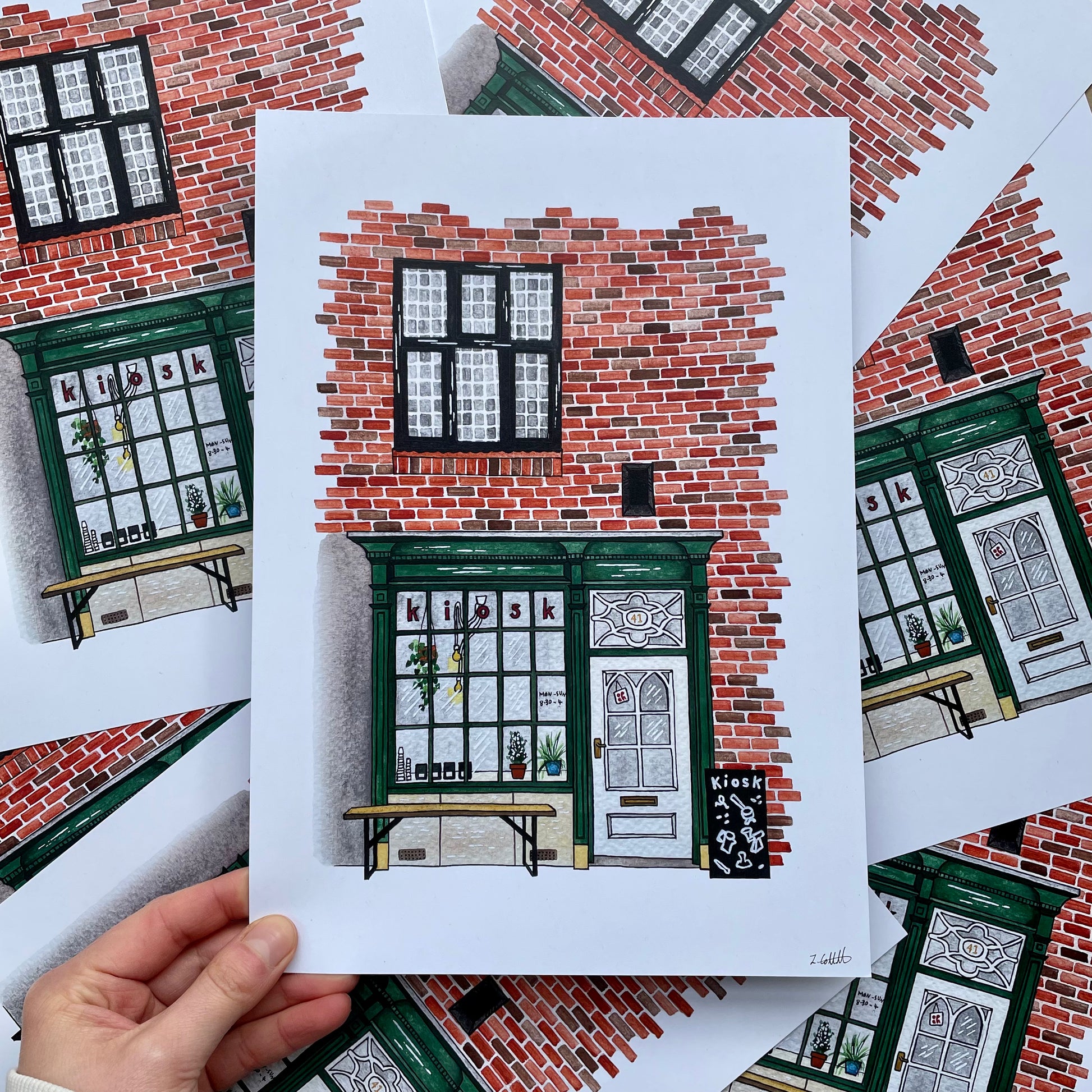 Hand holding a print of a brick building with green doors and windows.