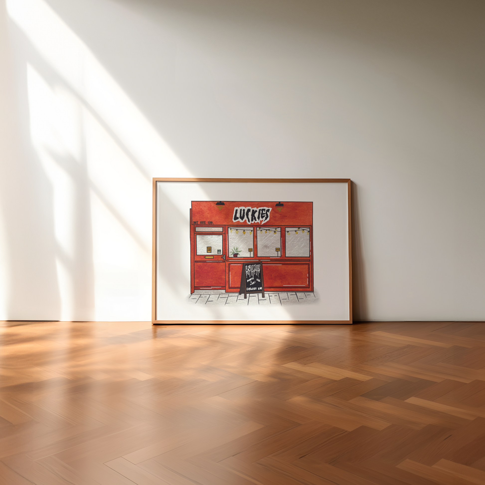 A framed art print depicting a red 'Luckies bar', leaning against a minimalist white wall on a polished wooden herringbone floor with soft sunlight casting shadows.