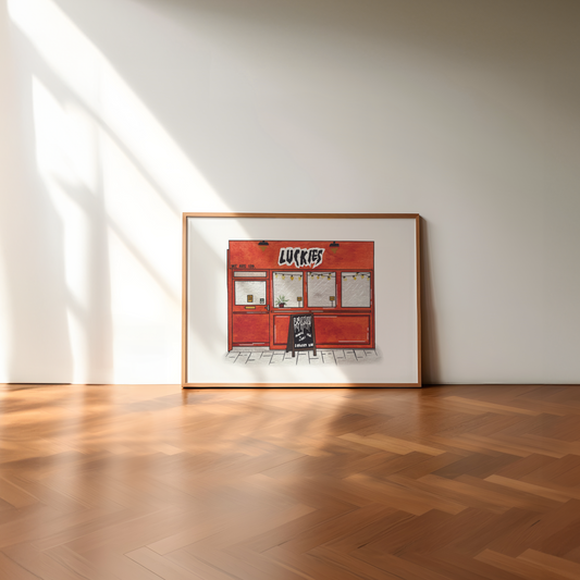A framed art print depicting a red 'Luckies bar', leaning against a minimalist white wall on a polished wooden herringbone floor with soft sunlight casting shadows.