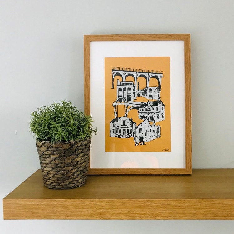 Framed artwork of ouseburn pubs print on a wooden shelf with a plant